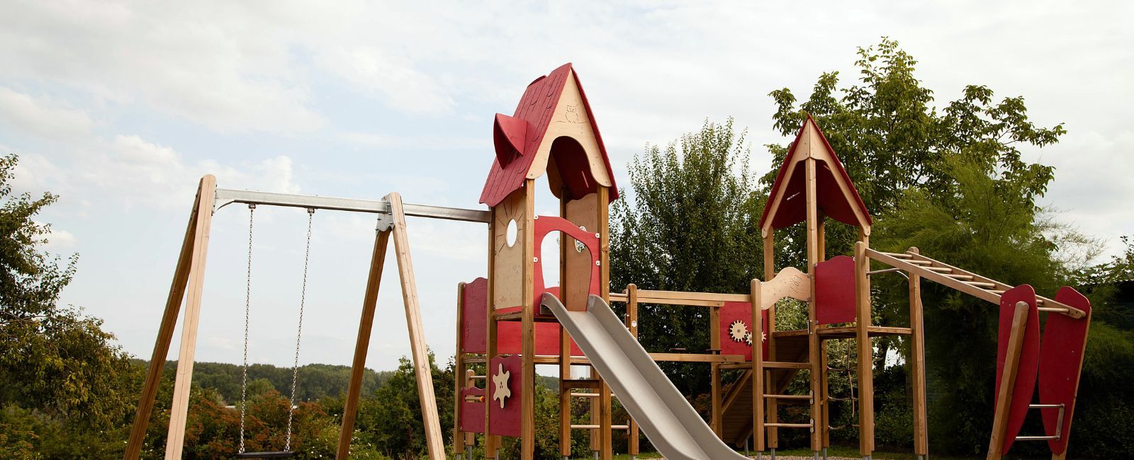 Image of swings, with wooden poles, a wooden playhouse with a red roof and a metal slide from the playhouse.